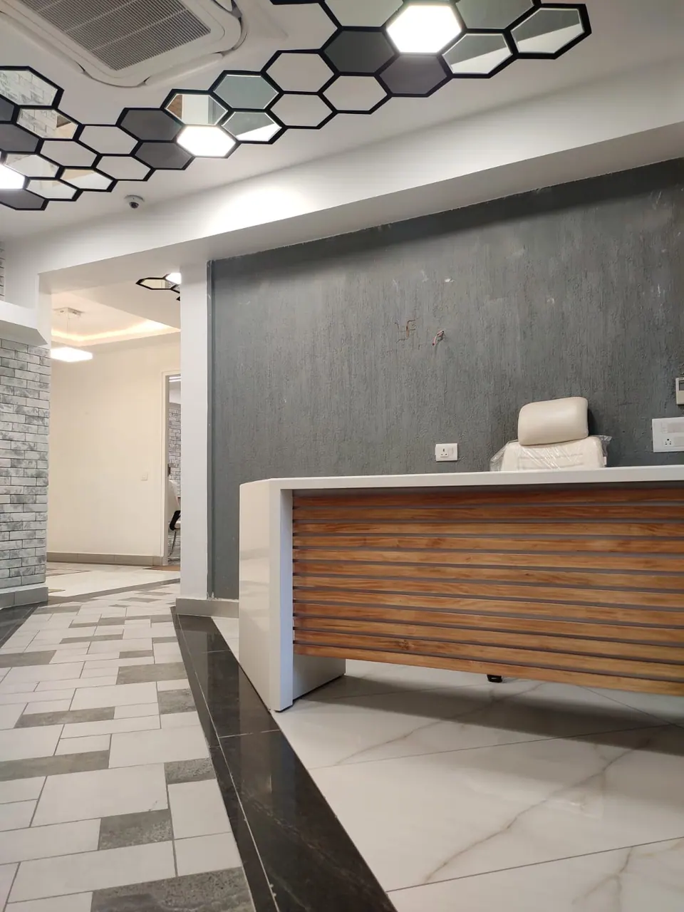 Reception counter in timber and white before a grey textured wall, a corridor with a brick-effect wall beyond