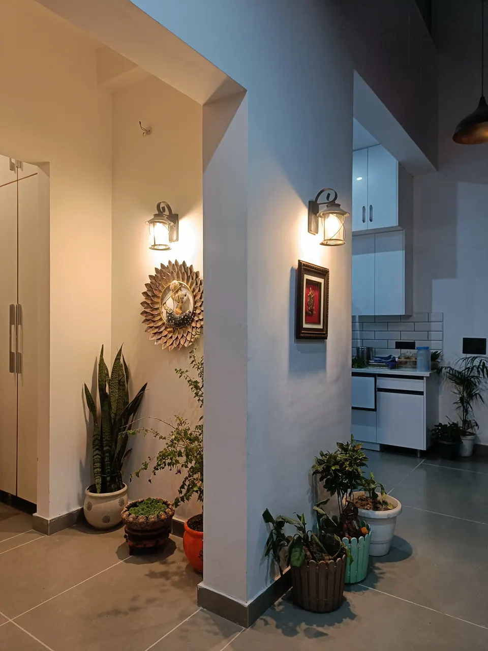 Entrance lobby with white walls, wall lanterns, a sunburst mirror and potted plants, the kitchen beyond