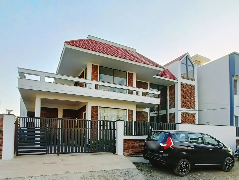 Two-storey house with a red tiled roof, white bands, brick jali panels and a glazed gable, a car parked by the gate