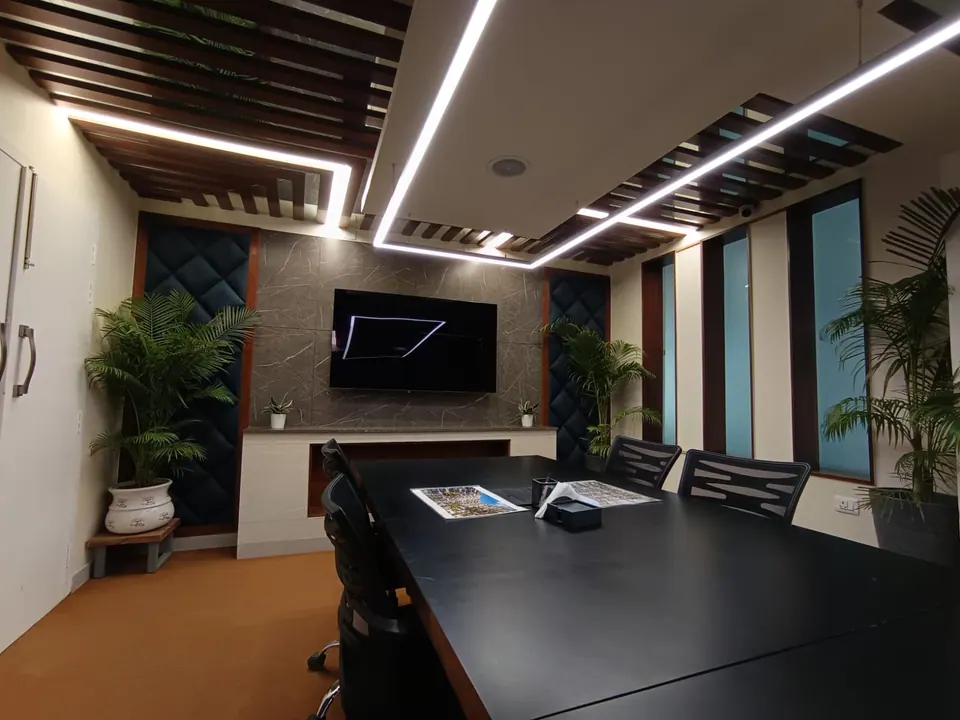 Conference room under a timber-slat ceiling with linear LED lights, a black table, mesh chairs and palms