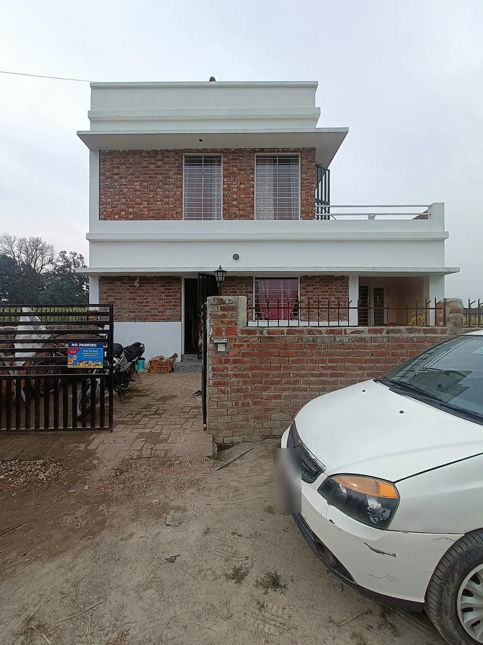 Two-storey brick house with white bands and a roof terrace behind a brick boundary wall, a car parked at the gate