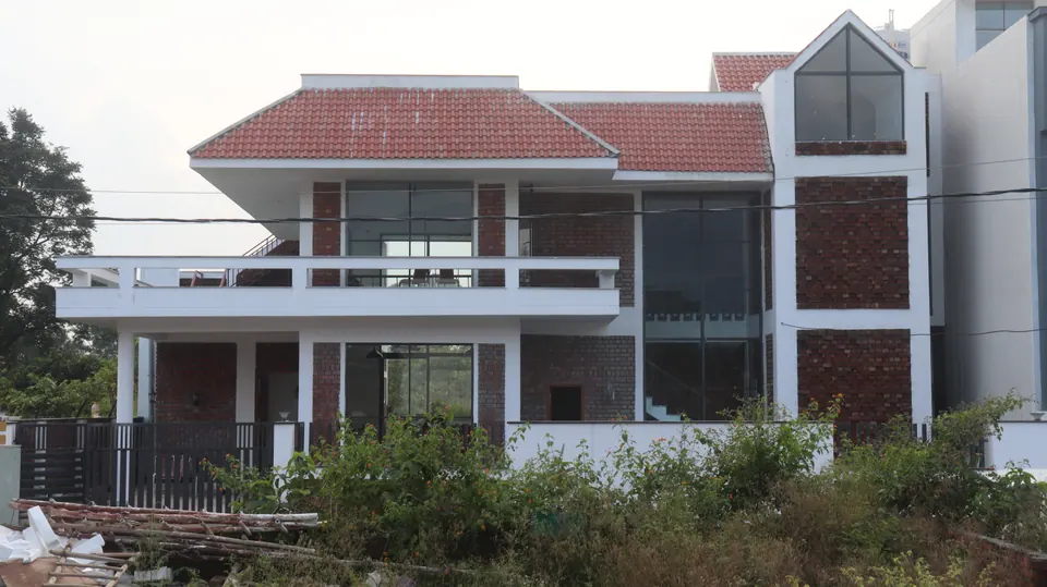Rear of the house over overgrown ground: a long balcony with white railings, brick walls, a red tiled roof and a glazed gable