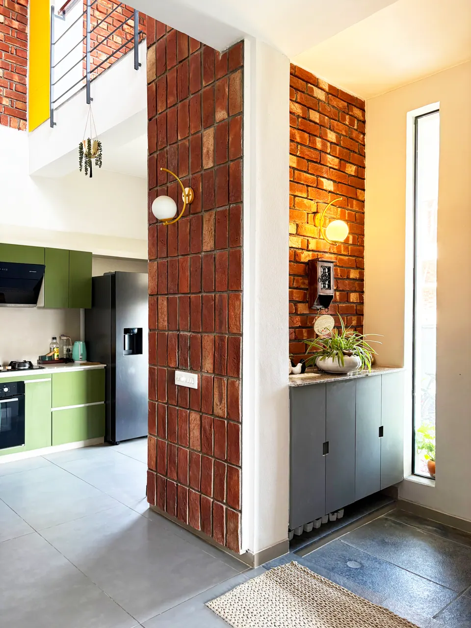 Double-height hall with a brick-clad column, a tall slit window, globe wall lights and the green kitchen beyond