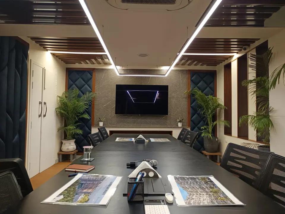 Conference table set with project brochures, facing a TV on a grey stone wall between upholstered panels and palms
