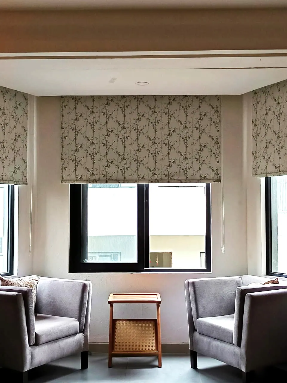 Two grey armchairs and a side table in a bay window with patterned Roman blinds