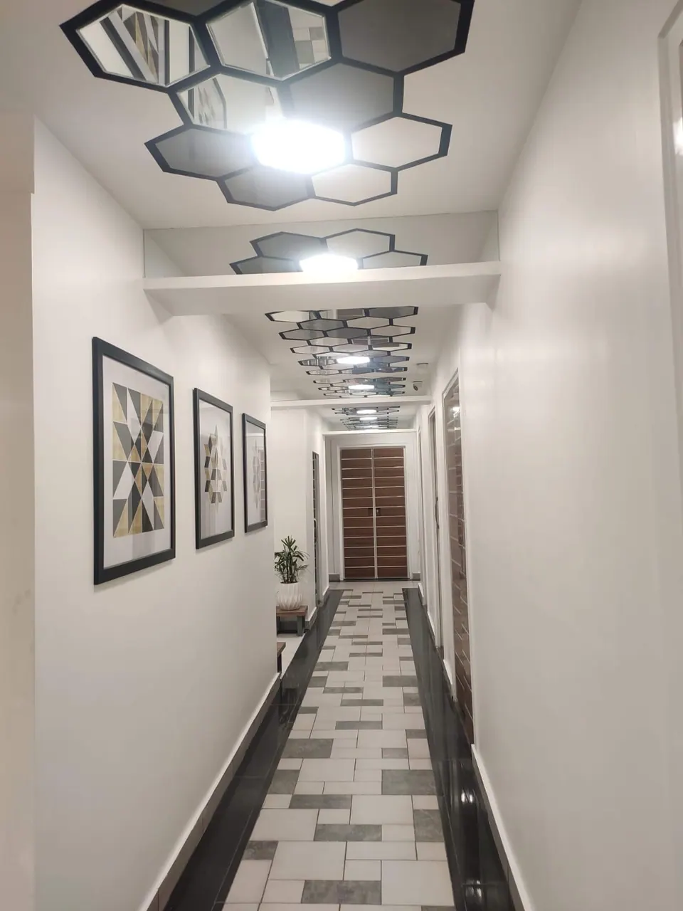 Narrow corridor under a run of hexagonal mirrored ceiling panels, with framed geometric prints and a patterned tile floor