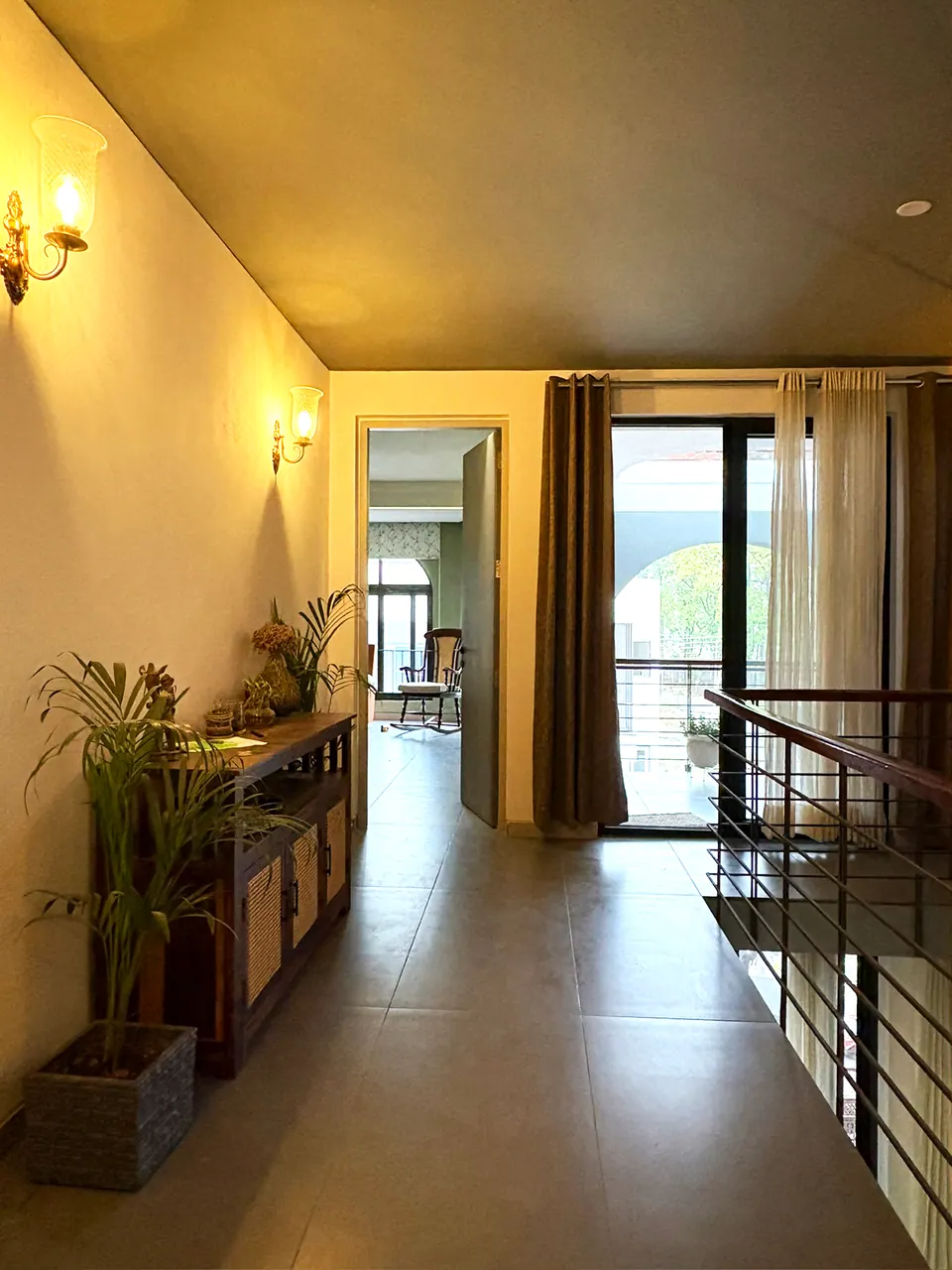 Upper hall with wall lamps, a timber console and plants, leading to a curtained balcony door