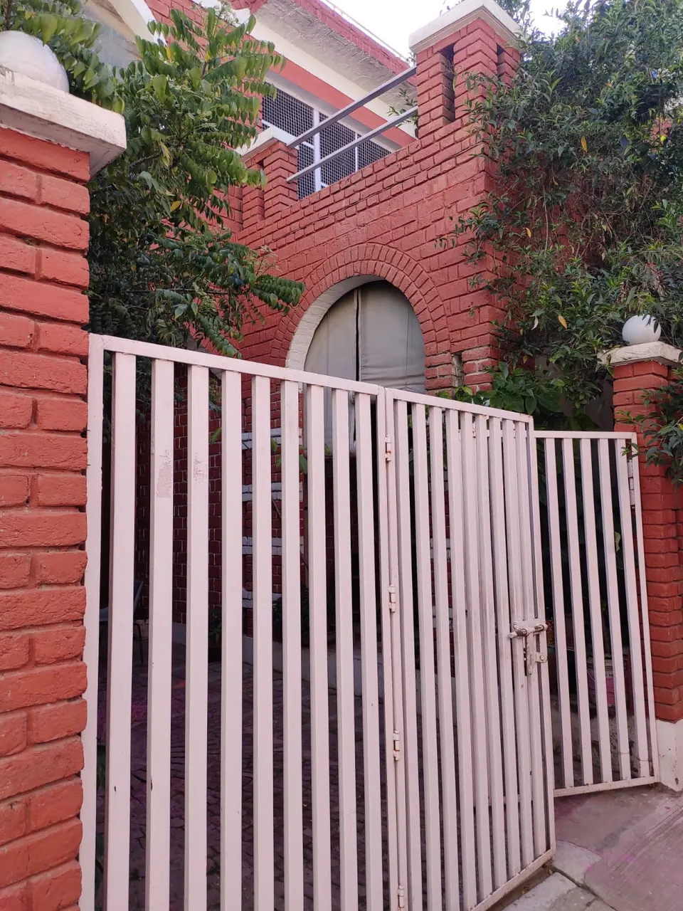 Entrance gate: white steel bars between red brick piers topped with globe lamps, with an arched brick opening and climbers behind
