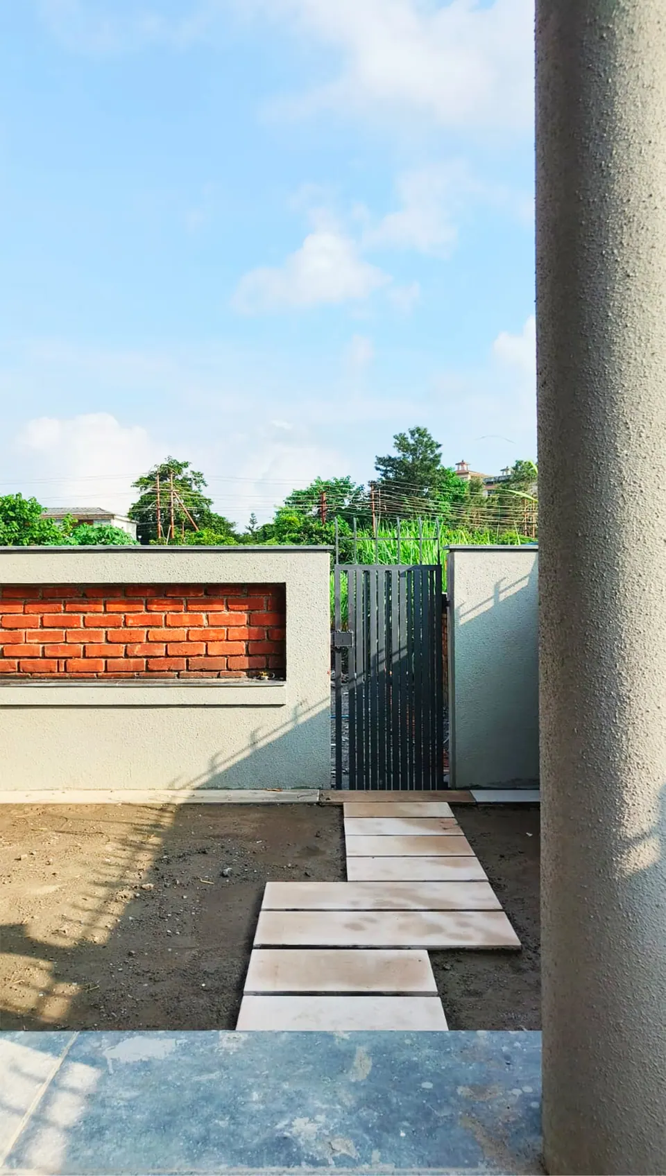 Stepping stones crossing bare ground to a gate in a rendered wall with a brick inset, a round column at the right