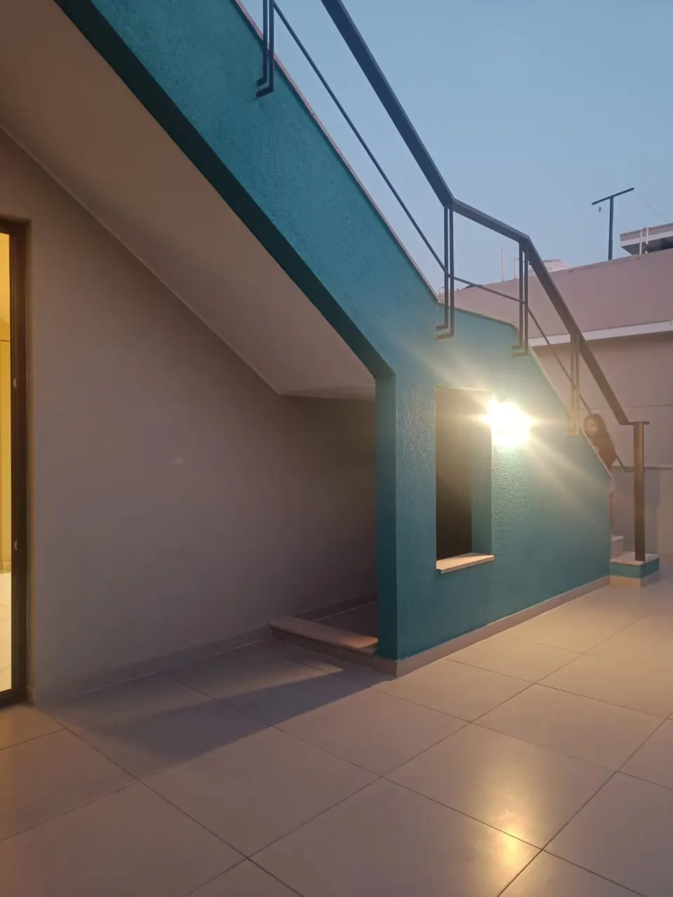Teal wall beneath the external stair, lit by a wall lamp, on the terrace at dusk