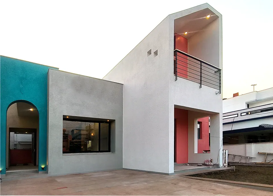 The house at dusk: white volumes, a tall inset balcony lit in coral, a teal wall with an arched entrance and a wide picture window