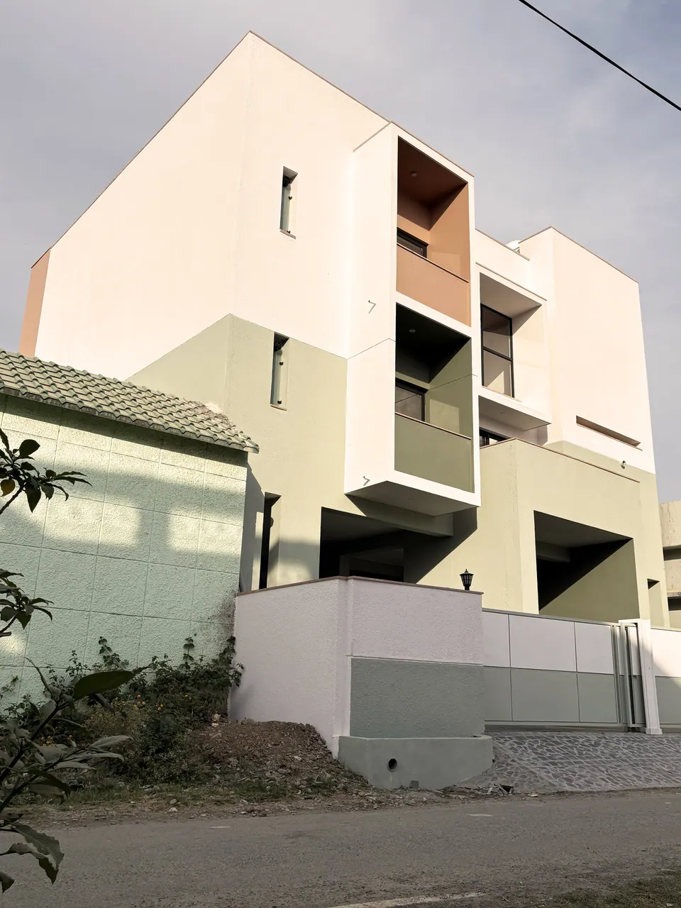 Corner of the white and sage three-storey house with a projecting balcony box and terracotta panel, rubble in the foreground