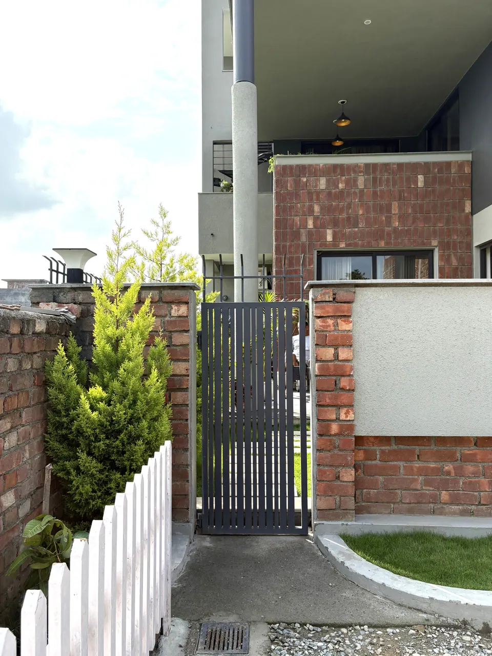 Side gate in a brick wall beside a white picket fence and a conifer, the brick-clad block above