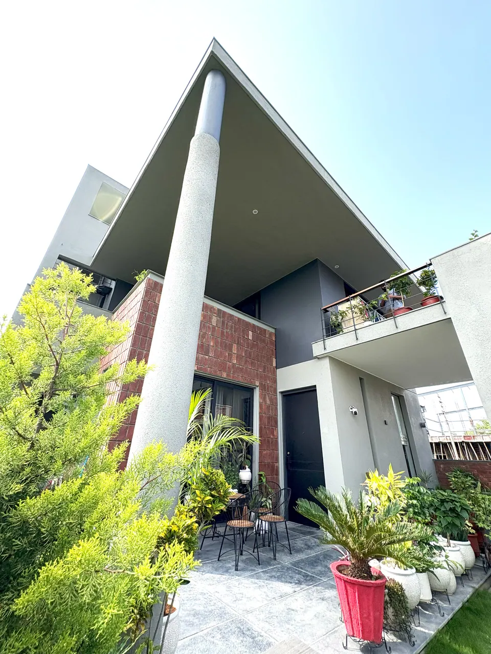 Upward view of the cantilevered roof on a round column above the brick-clad block, a planted patio with chairs below
