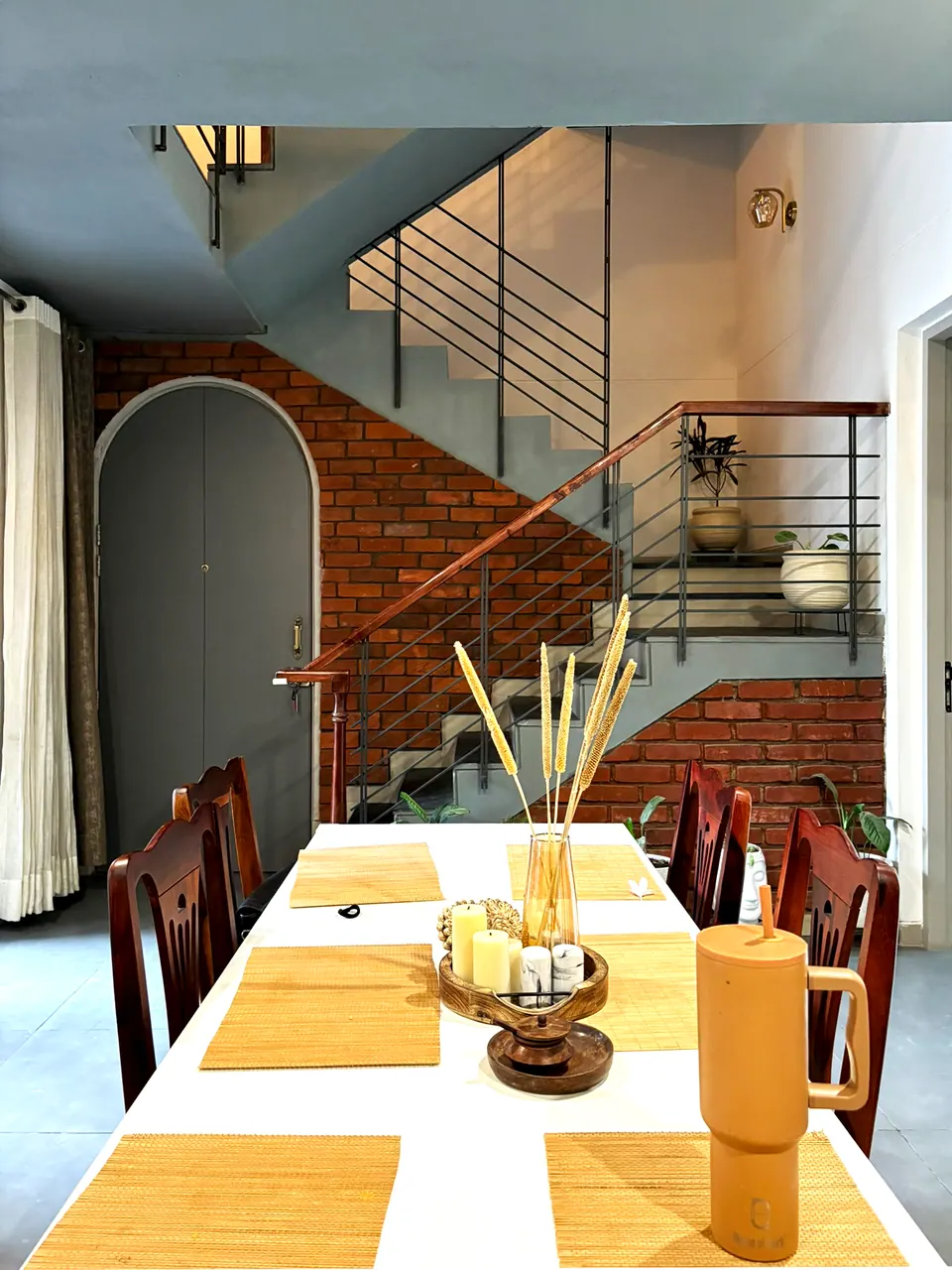 Dining table set with bamboo mats before a brick-walled stair with a black steel railing and an arched grey door