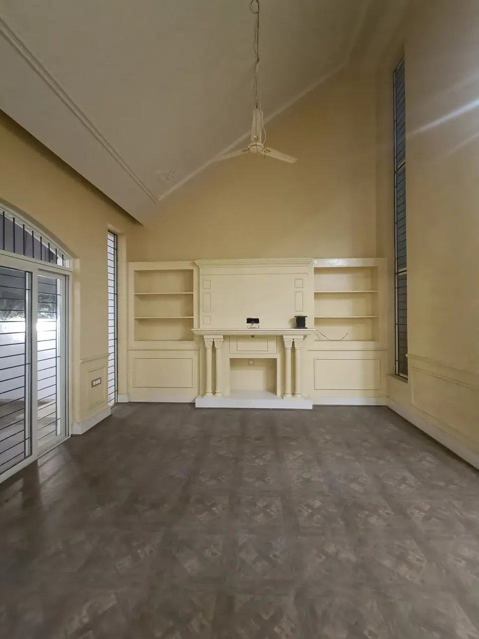 High-ceilinged room with a white fireplace between built-in shelves, tall slit windows and a ceiling fan