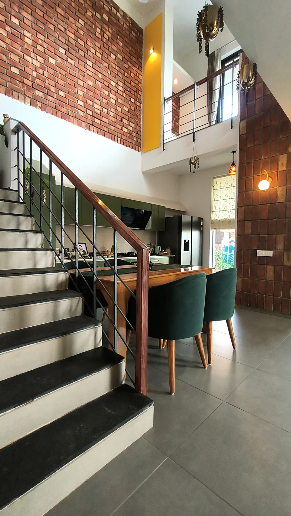 Stair with dark treads and a timber handrail rising past the dining table, the green kitchen and brick walls beyond