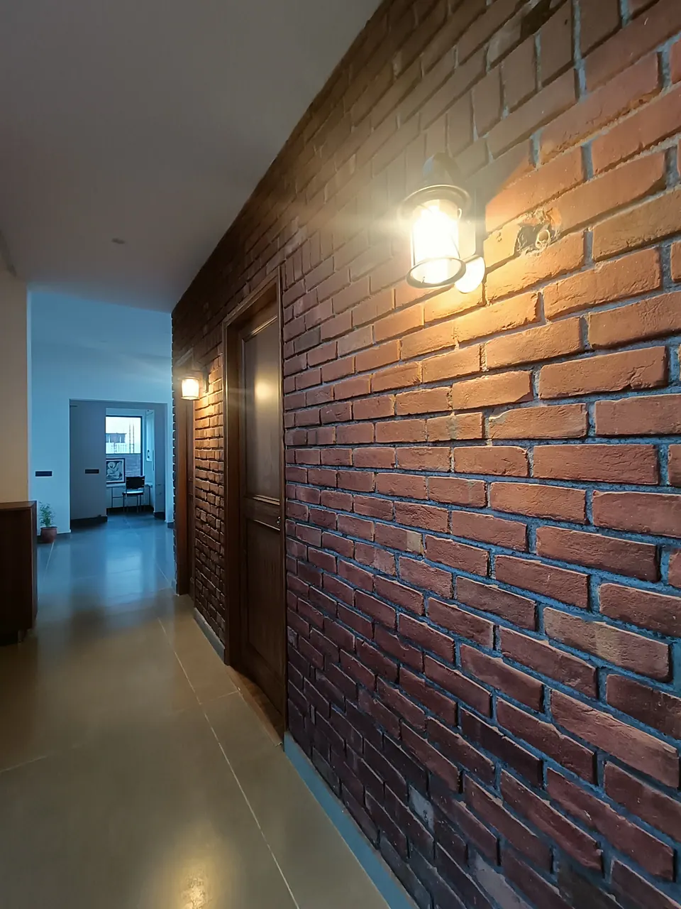 Corridor with an exposed brick wall and a lit wall lantern leading to a bright room