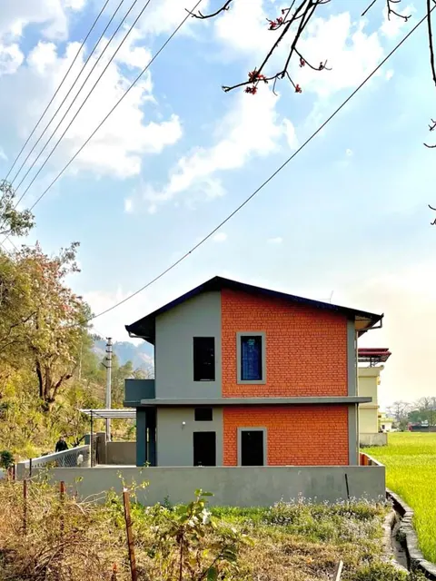 Two-storey gabled house in grey render and orange brick on a raised plinth beside a green field, wooded hills behind