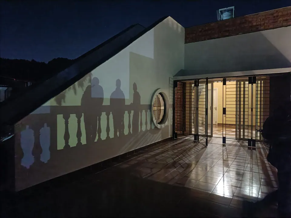 Roof terrace at night: lamplight throws the shadows of the balustrade and of three people onto a white wall beside a round window