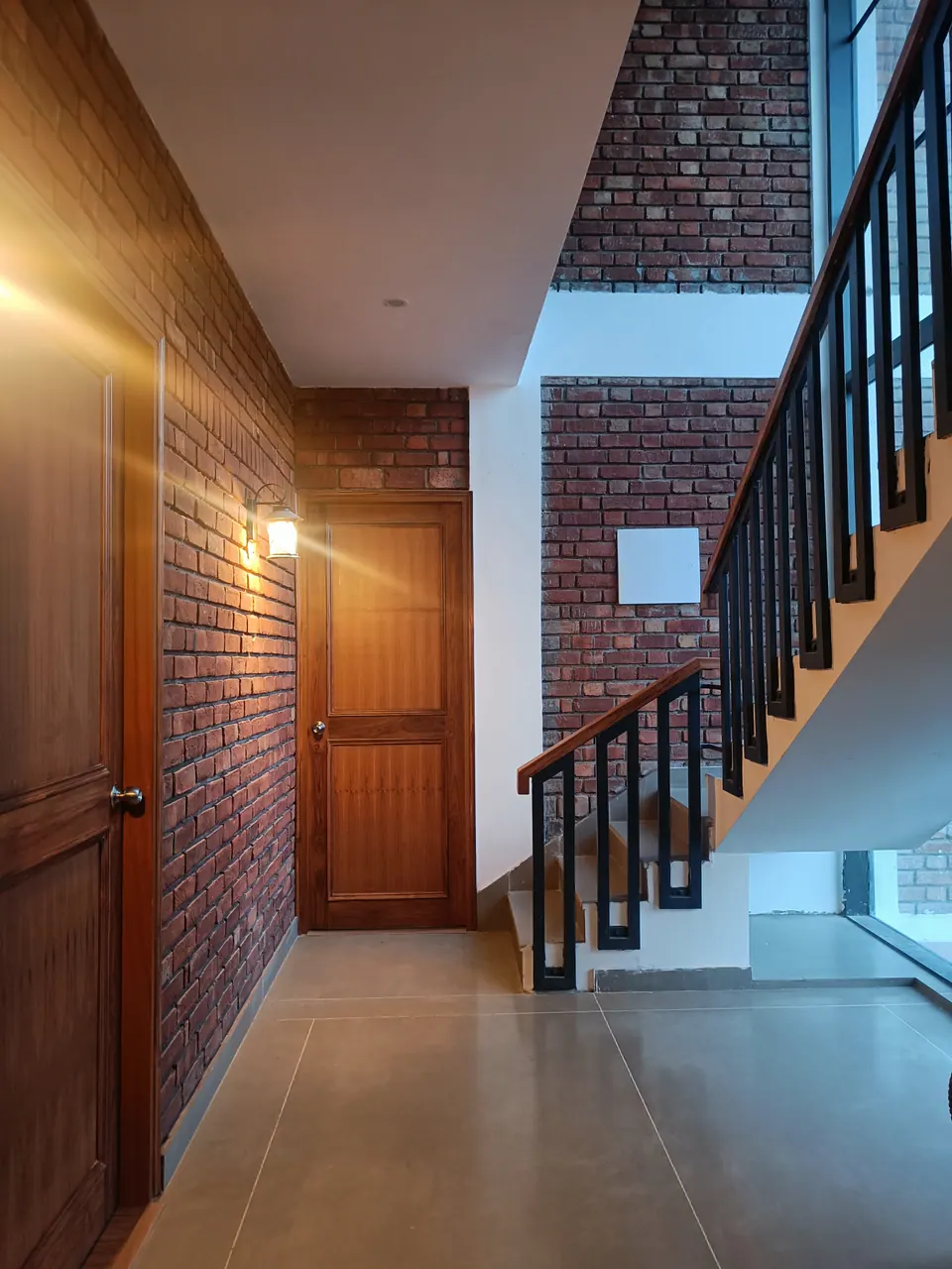Hall with an exposed brick wall, timber doors, a wall lantern and a stair with a black steel balustrade and timber treads