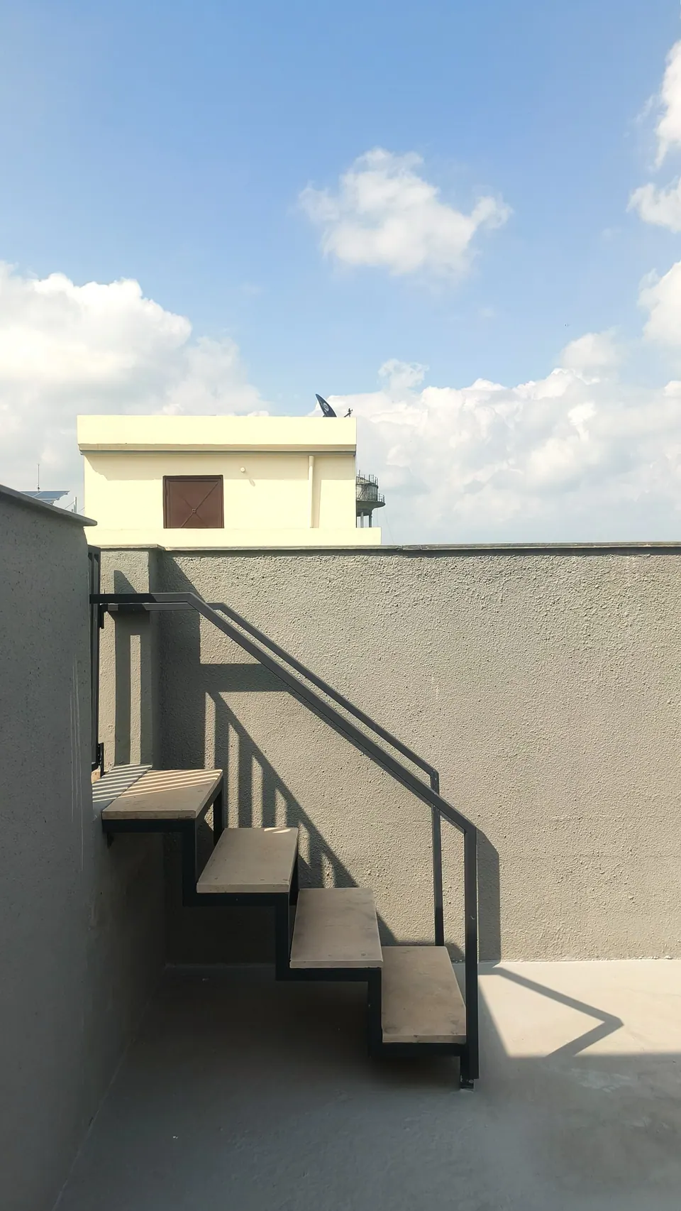 Steel stair with cantilevered grey treads rising along a grey parapet wall to the roof, under a blue sky