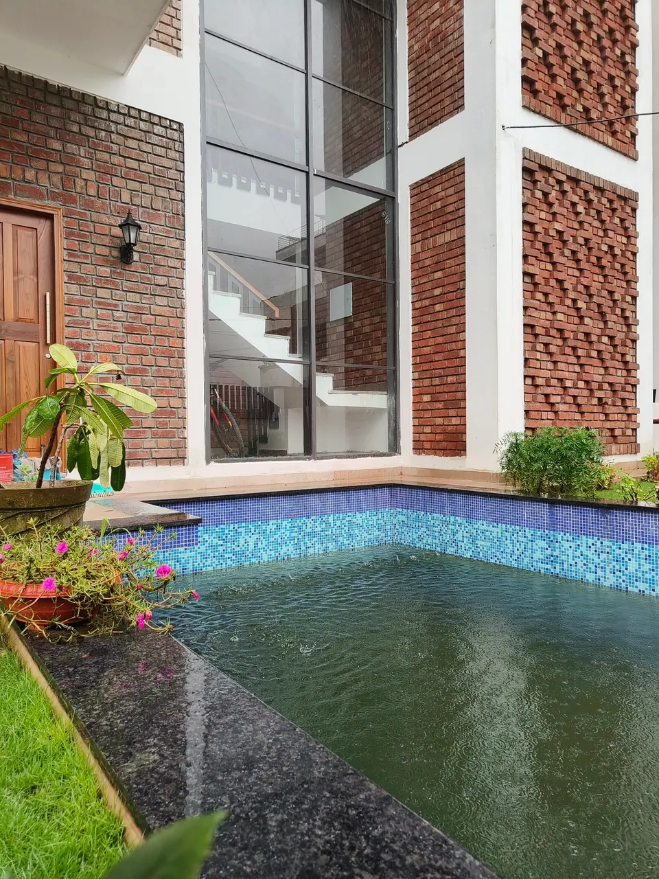 Pool with blue mosaic tiles and a black granite edge beside a brick wall with jali panels and full-height glazing
