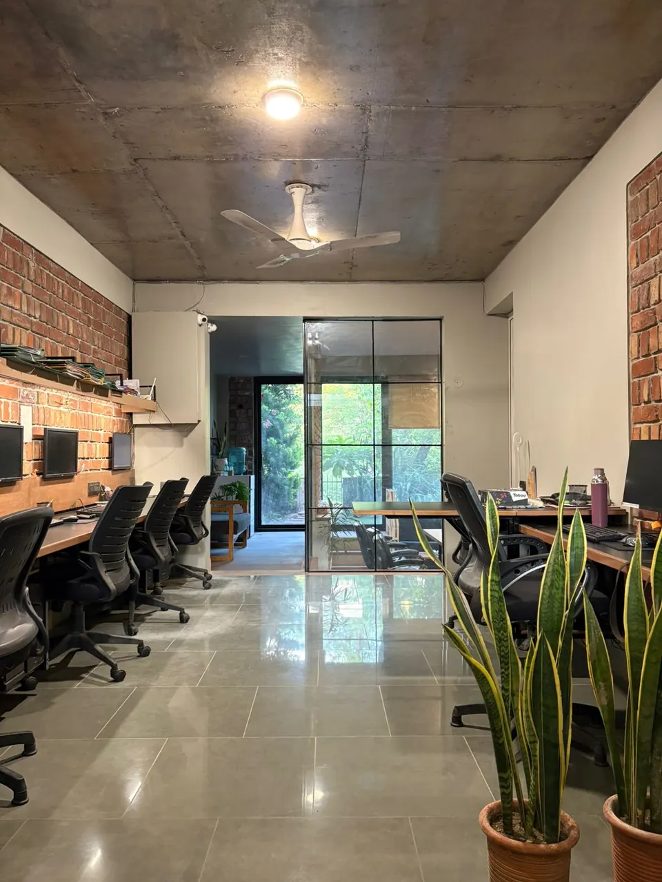 The studio: a row of workstations along a brick wall under an exposed concrete ceiling, potted snake plants, and a glazed door to the garden at the far end