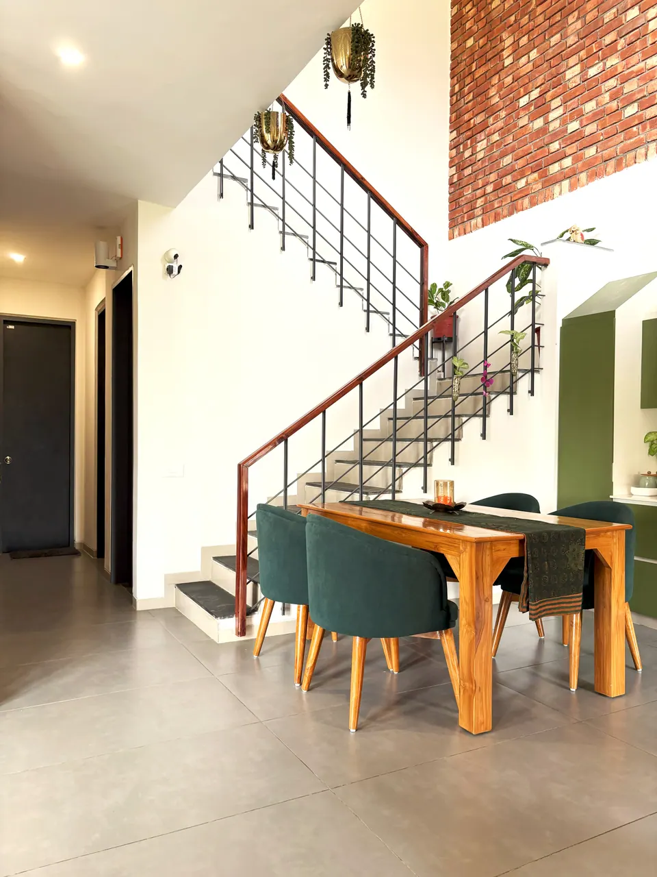 Dining area with a teak table and green velvet chairs beneath a stair with a timber handrail, a brick wall above