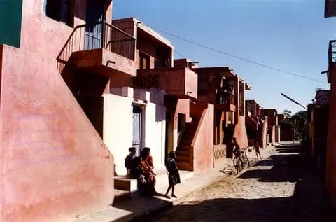 Pink-plastered row houses with stairs and terraces along a sunlit lane, with residents sitting by a doorway and a cyclist further down