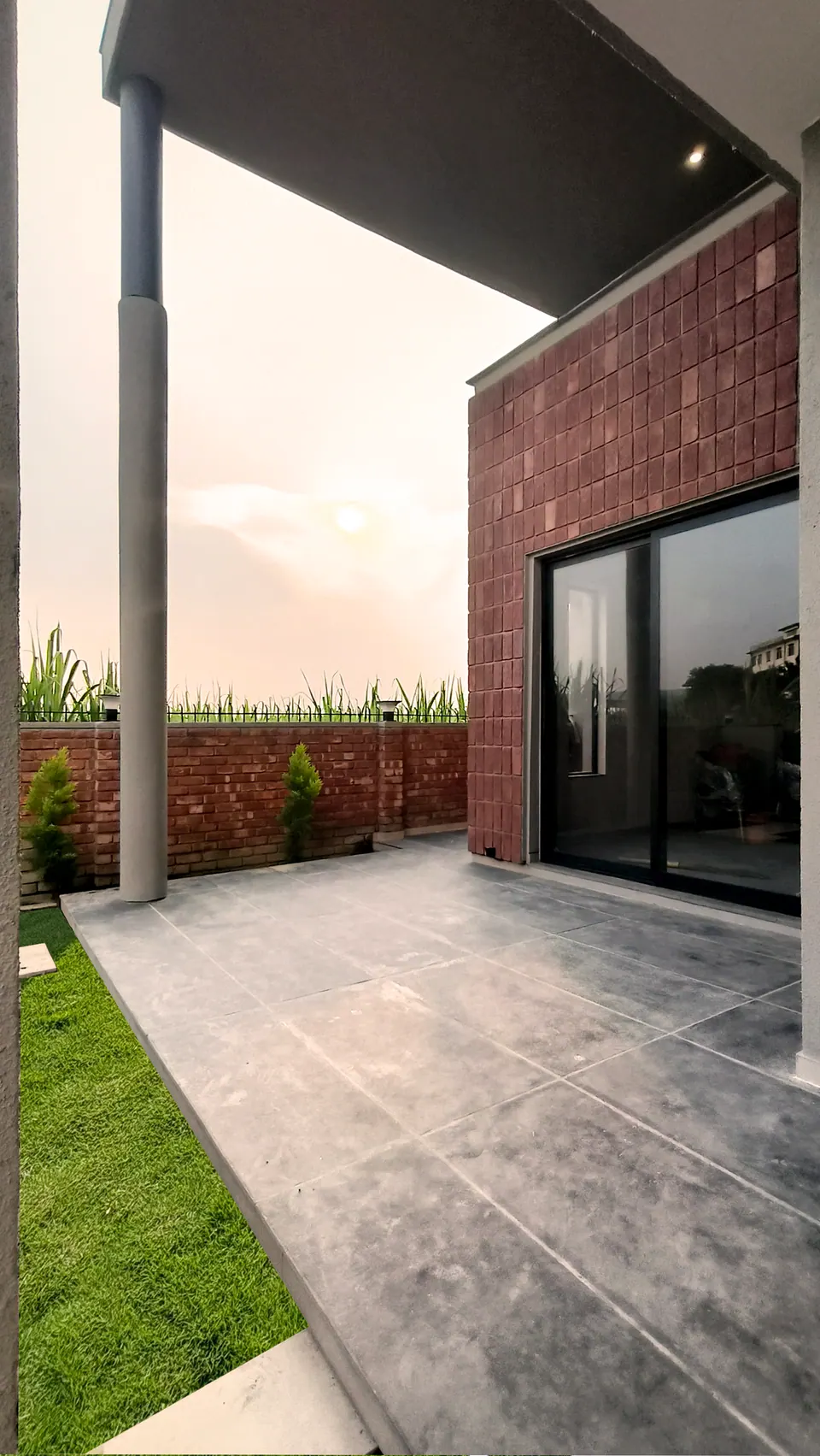 Covered terrace under the cantilevered roof beside a brick-clad wall with a sliding glazed door, a lawn edge in front