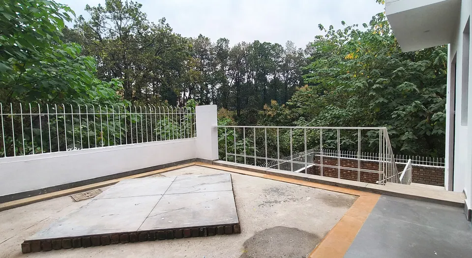 Paved terrace with a white parapet and railings and a raised brick-edged platform, trees beyond