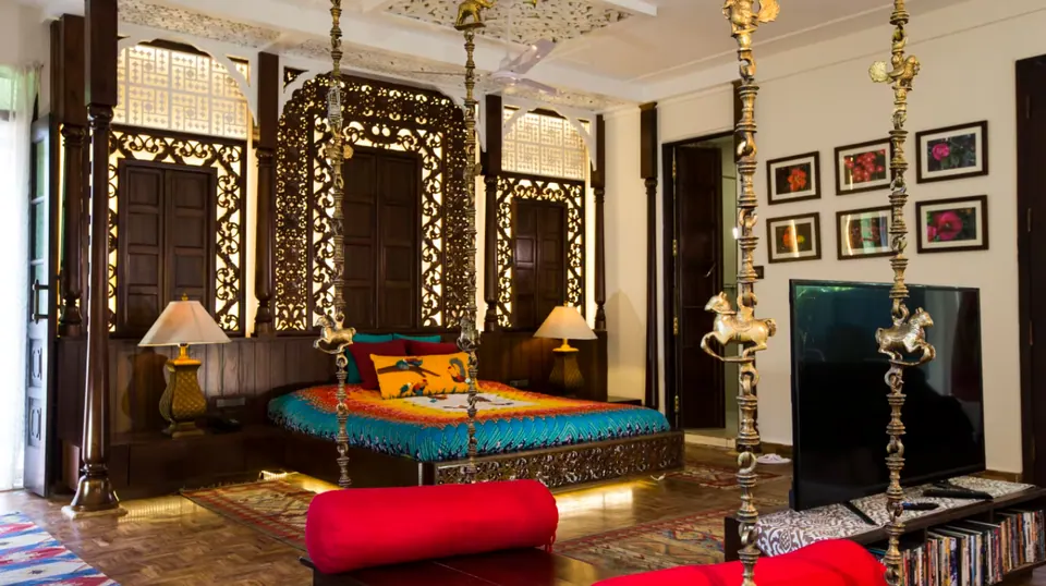 Bedroom with carved timber screens behind a bed with a bright cover, a swing on brass chains, framed pictures and kilim rugs