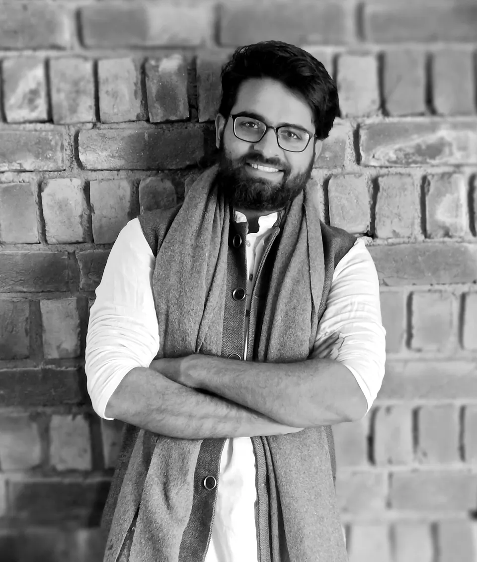 Black-and-white portrait of Surya, arms folded, wearing glasses and a scarf, in front of a brick wall