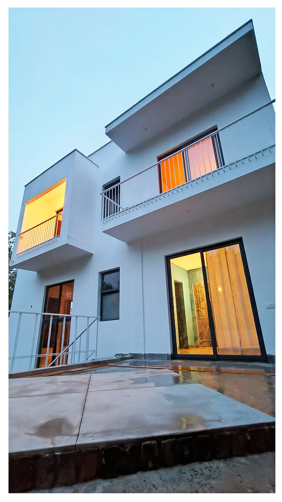 White-rendered elevation at dusk, lit windows and a projecting balcony above a wet paved terrace