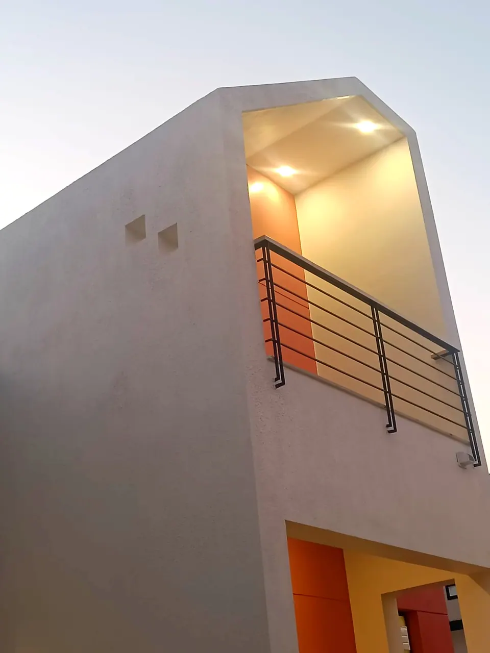 Tall white volume with a deep inset balcony, its black steel railing and warm-lit coral recess against the evening sky