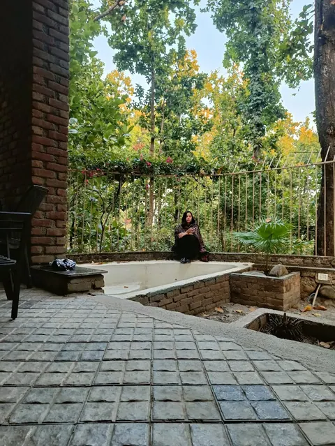 A woman seated on the edge of an empty sunken pool in a stone-paved courtyard, beside a brick pillar and a railing, with tall trees beyond