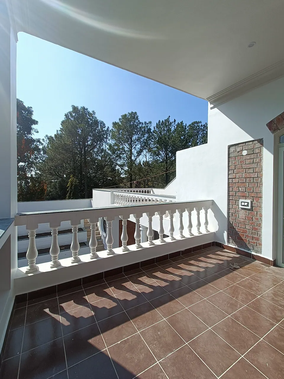 Balcony with a white balustrade casting shadows on terracotta tiles, with pine trees and the white terraces beyond