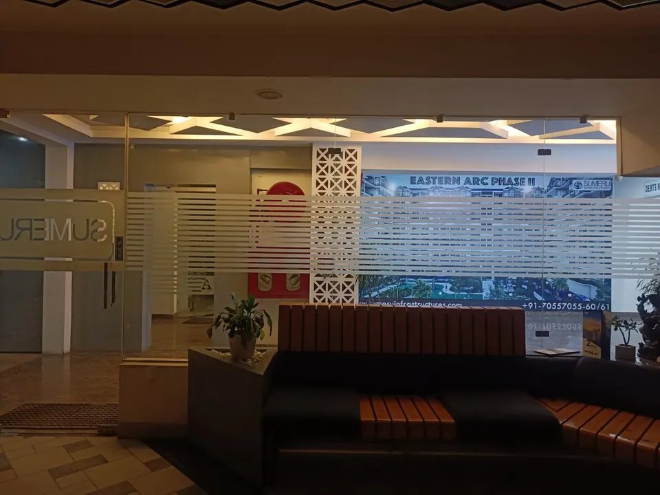 Glass entrance at night with a frosted "SUMERU" sign, a timber-slatted sofa and a project banner on the wall beyond