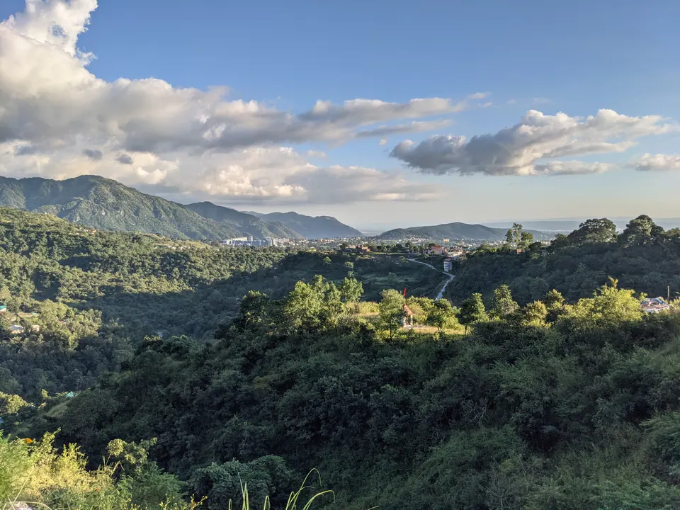 Forested hills in Uttarakhand falling to a town in the valley, with a mountain ridge beyond under scattered clouds