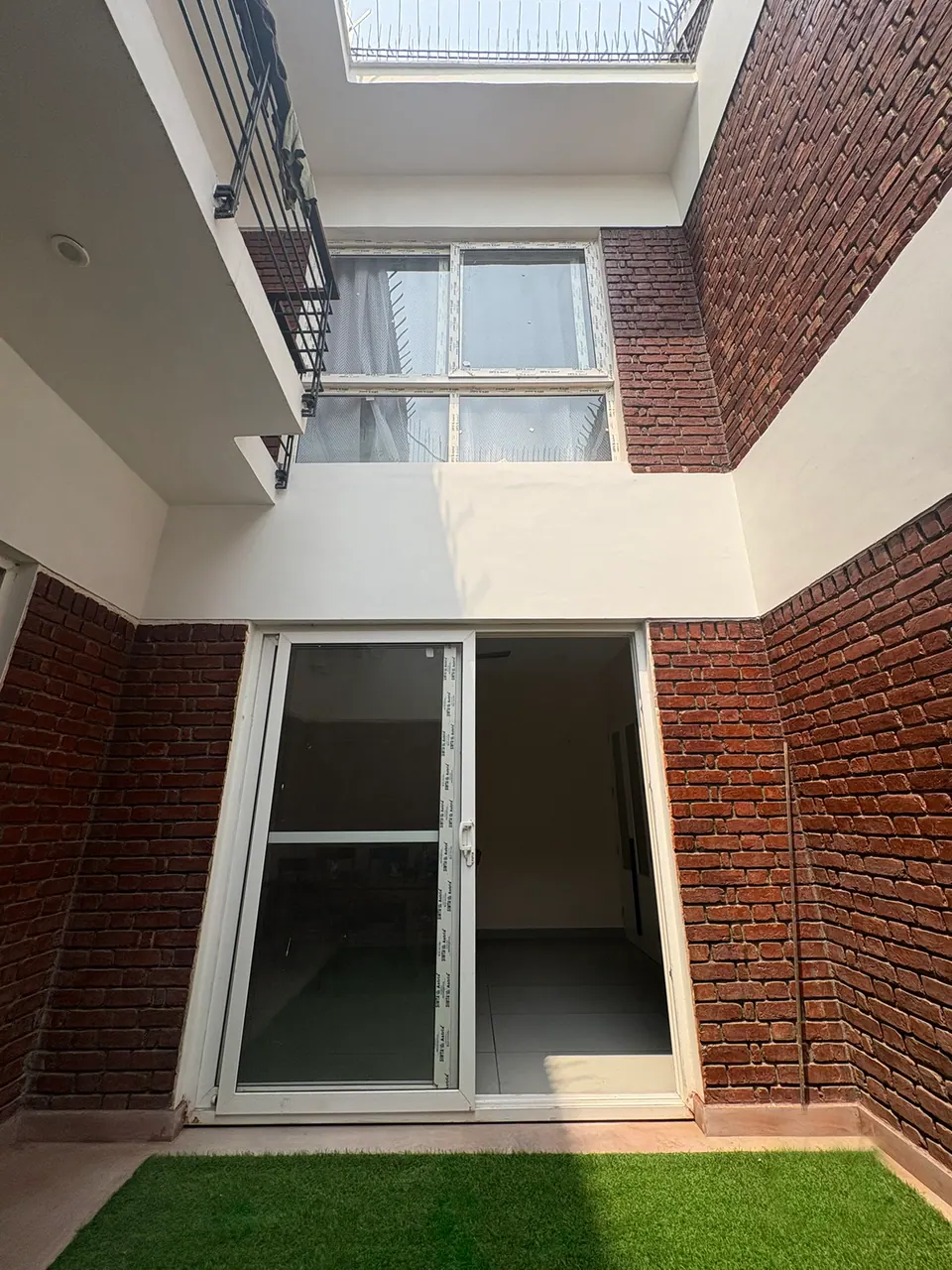 Courtyard with artificial turf before a white-framed sliding glass door, between brick walls, with a window above