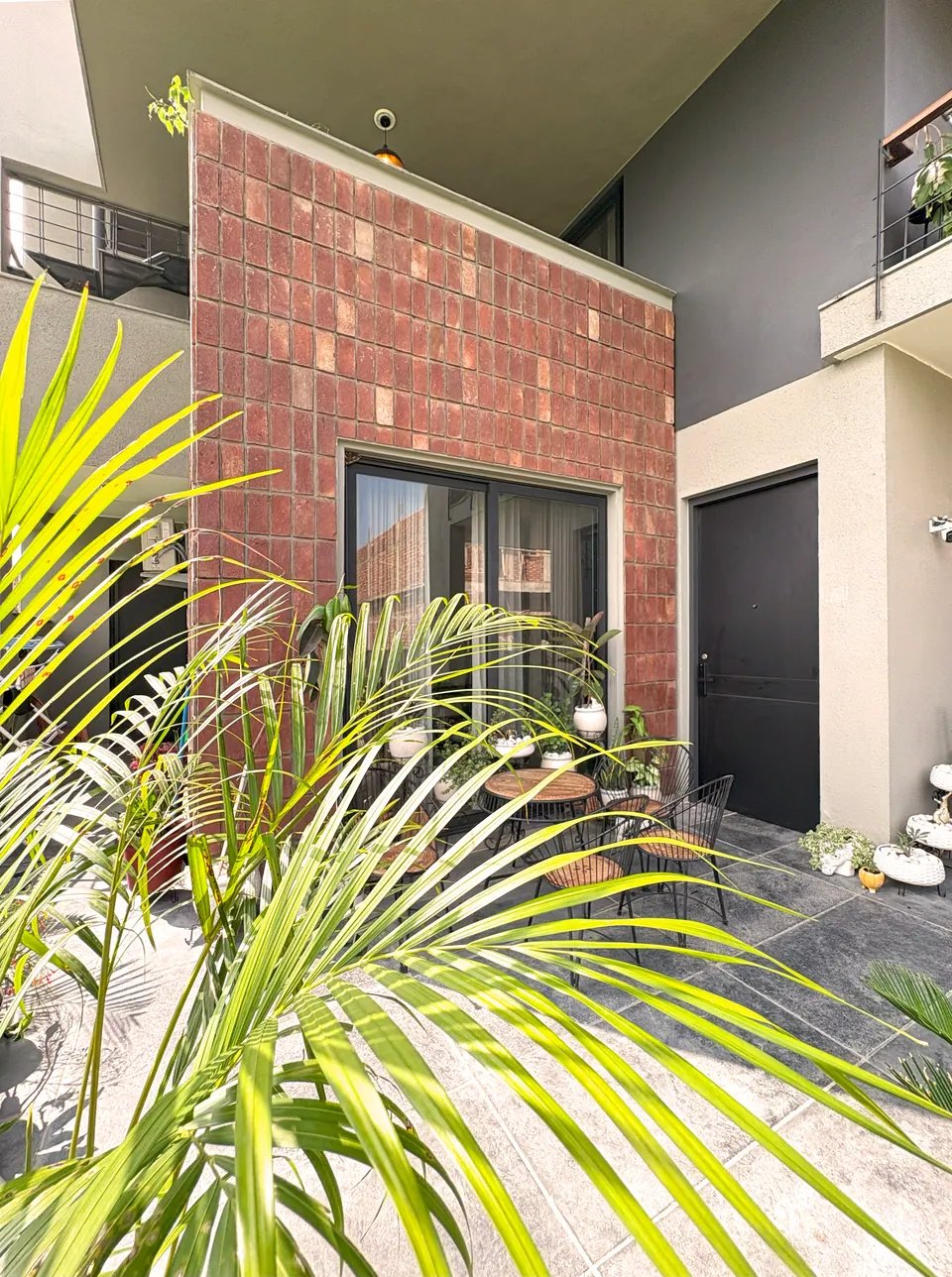 Brick-clad block with a glazed door beside a dark entrance door, a patio with a cafe table behind palm fronds