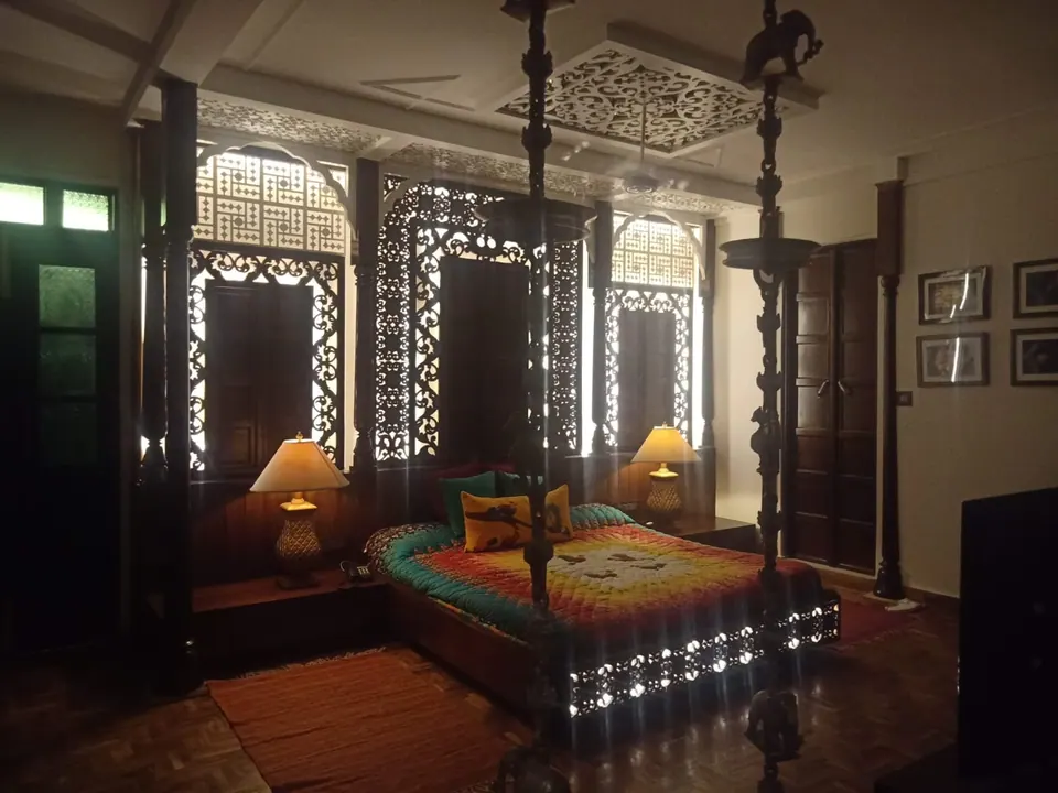 Dim bedroom with carved timber lattice screens over the windows, a bed with a bright patchwork cover, table lamps and a swing chain