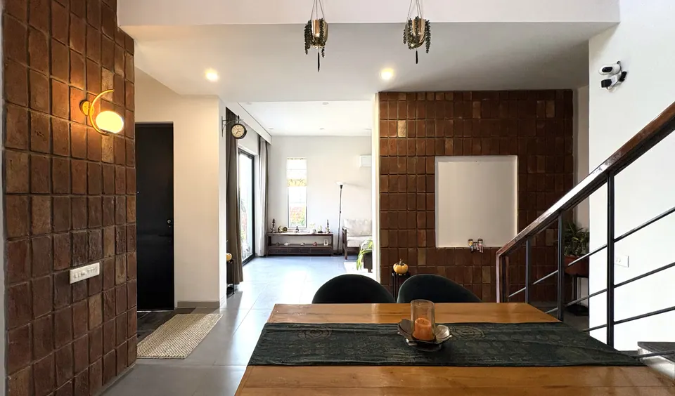 Dining table with green chairs before a brick-tile wall and globe lights, the living room beyond