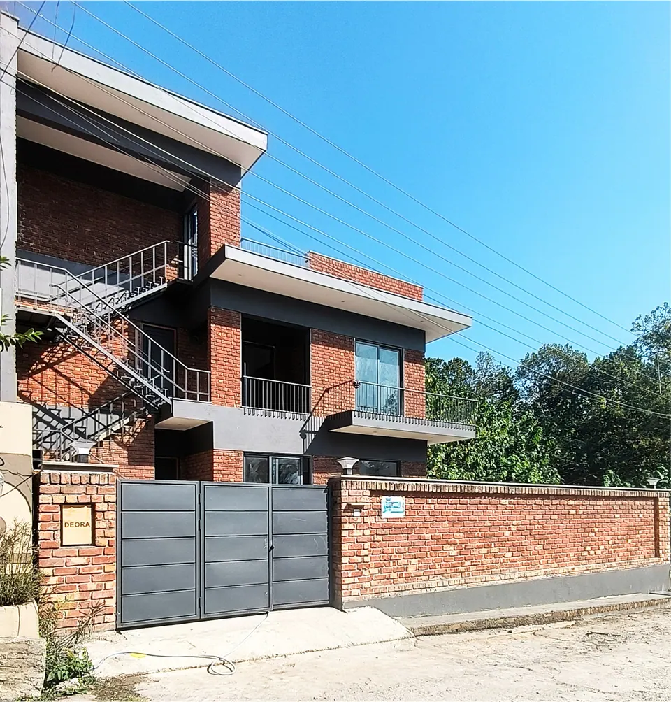 Street front with a grey steel gate in a brick boundary wall and the two-storey brick house rising behind
