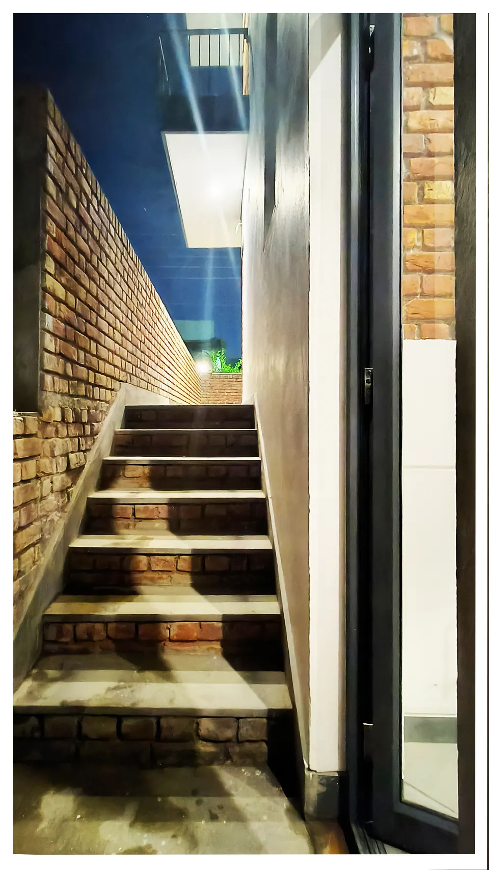 Narrow stair between a brick wall and a white-rendered wall at night, lit from above