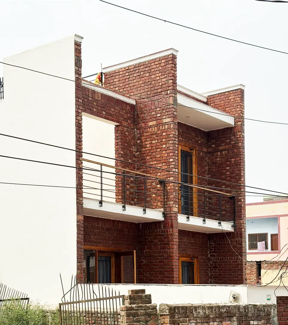 Street view of the two-storey house in exposed red brick, with cantilevered white slabs, a railed balcony and a framed roof opening