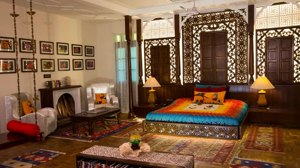 Bedroom with carved timber lattice panels behind the bed, a swing with a red bolster, a fireplace, white armchairs and rugs