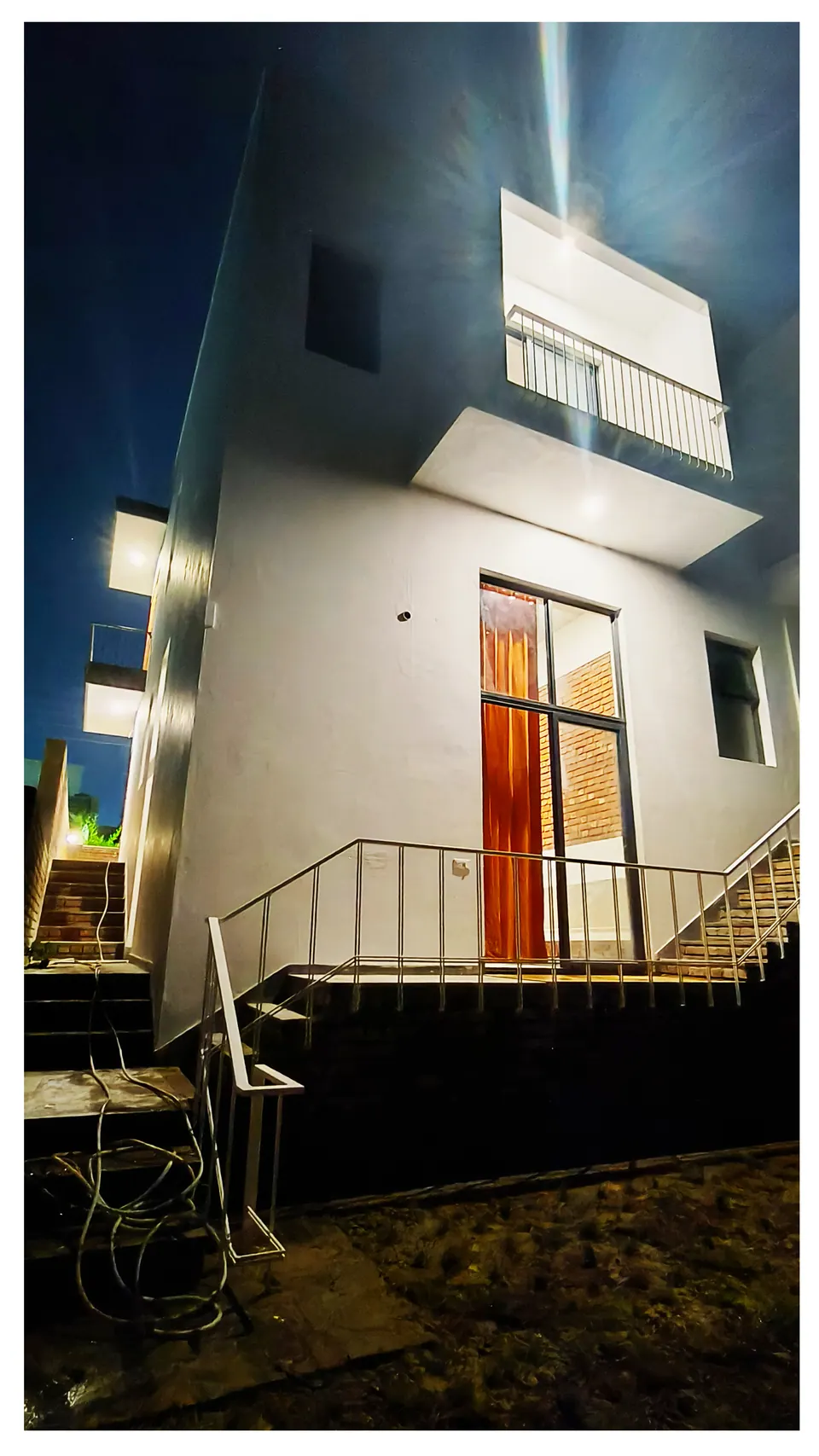 Night view of a white wall with a timber door, a projecting balcony light and steps with a white railing
