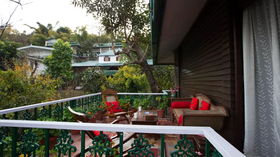 Balcony with a green cast-iron railing over a garden with cane seating and red cushions, hillside houses beyond